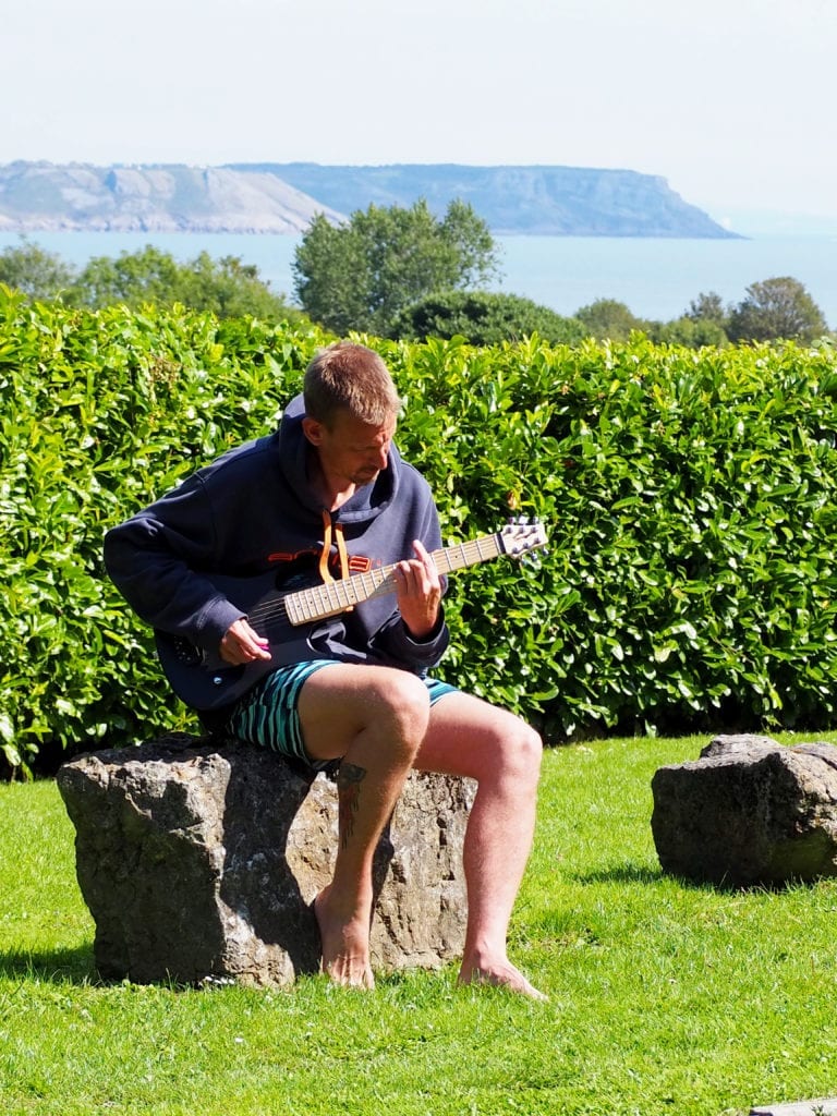 5 Reasons Why a Guitarist Needs a Snap Dragon in their Life - Ben sitting on a rock playing the guitar with grass behind him and the sea in the distance