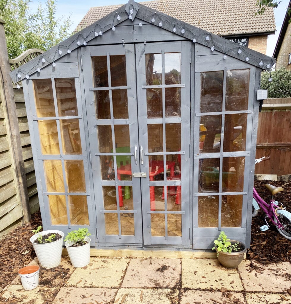 Billy Oh 6 x 8 Summerhouse Review - picture of a wooden summer house with square windows and doors at the front. Painted slate grey and fairy lights hanging on the front.