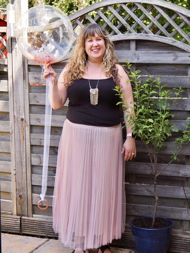 A picture of me in a black top and pink skirt holding a balloon with happy 40th on it in the garden