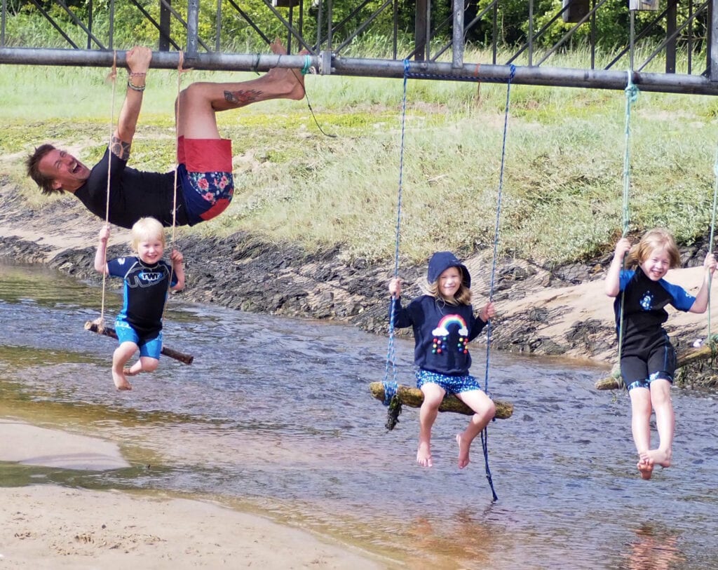 Three children on rope swings hanging off a bridge with their Dad photo bombing behind hanging off of the bridge
