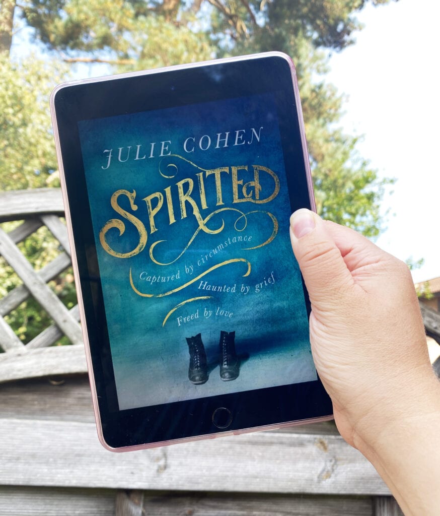 Book Review: Spirited by Julie Cohen. Picture of an iPad Mini being held up in front of some trees with the book cover for Spirited displayed. 